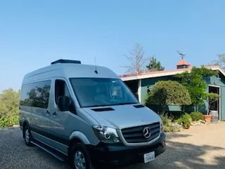 A silver Mercedes Sprinter van parked outside a farmhouse under a clear sky